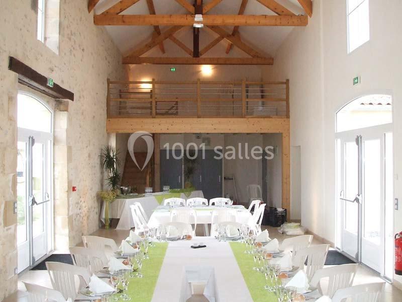 Salle de réception lumineuse avec poutres apparentes, tables dressées et mezzanine en bois.