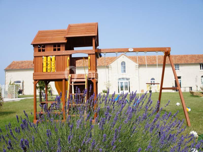Structure en bois avec toboggan et balançoire dans un jardin, devant un bâtiment en pierre et des lavandes au premier plan.