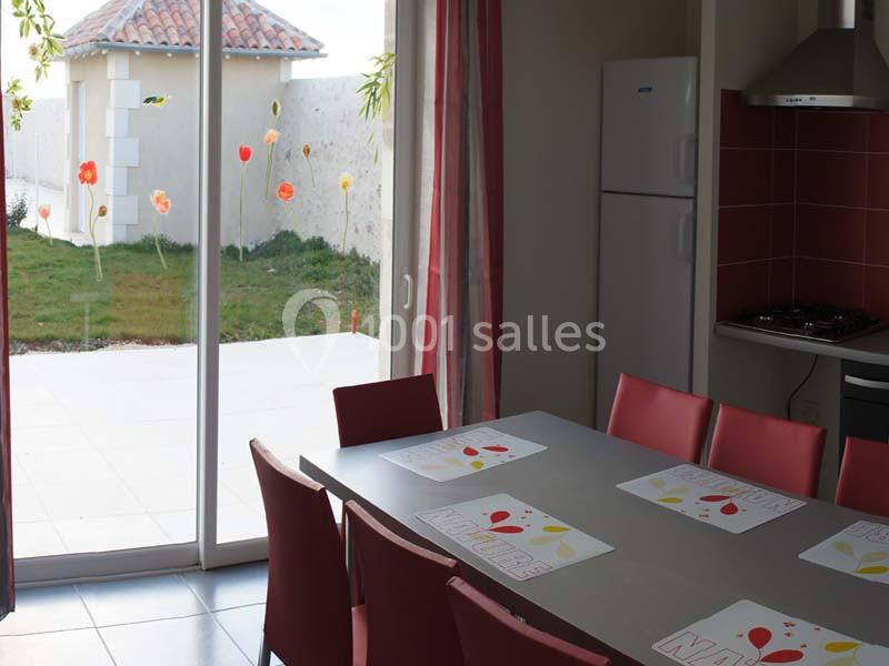 Salle à manger lumineuse avec table grise, chaises rouges, et vue sur une terrasse et un jardin fleuri par une baie vitrée.
