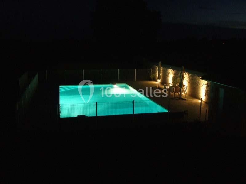 Piscine éclairée de nuit, entourée d'une clôture et de chaises longues sous des parasols.