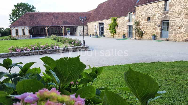 Cour d'une ferme rénovée avec bâtiments en pierre, jardin fleuri au premier plan et pelouse bien entretenue.