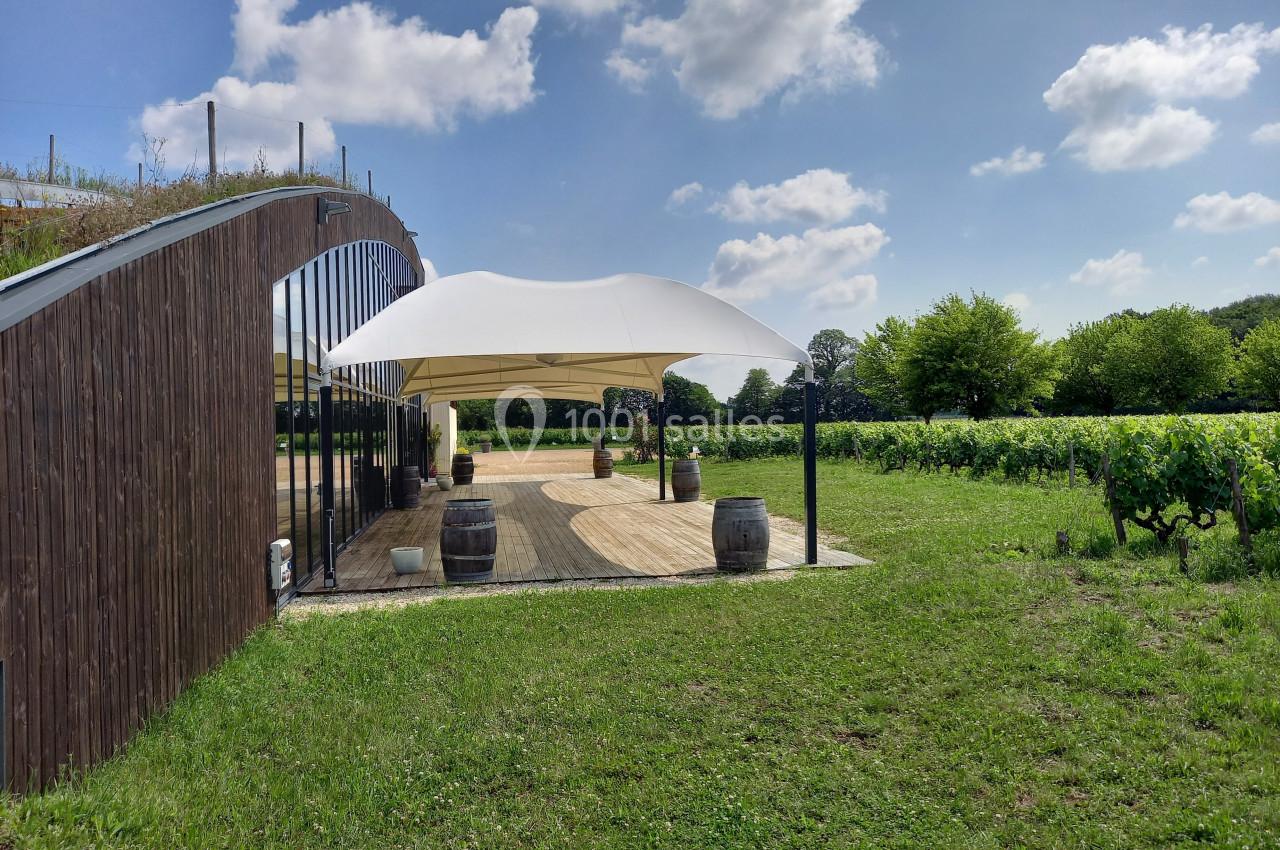 Terrasse en bois avec tonnelle blanche, entourée de vignes et d'arbres sous un ciel partiellement nuageux.