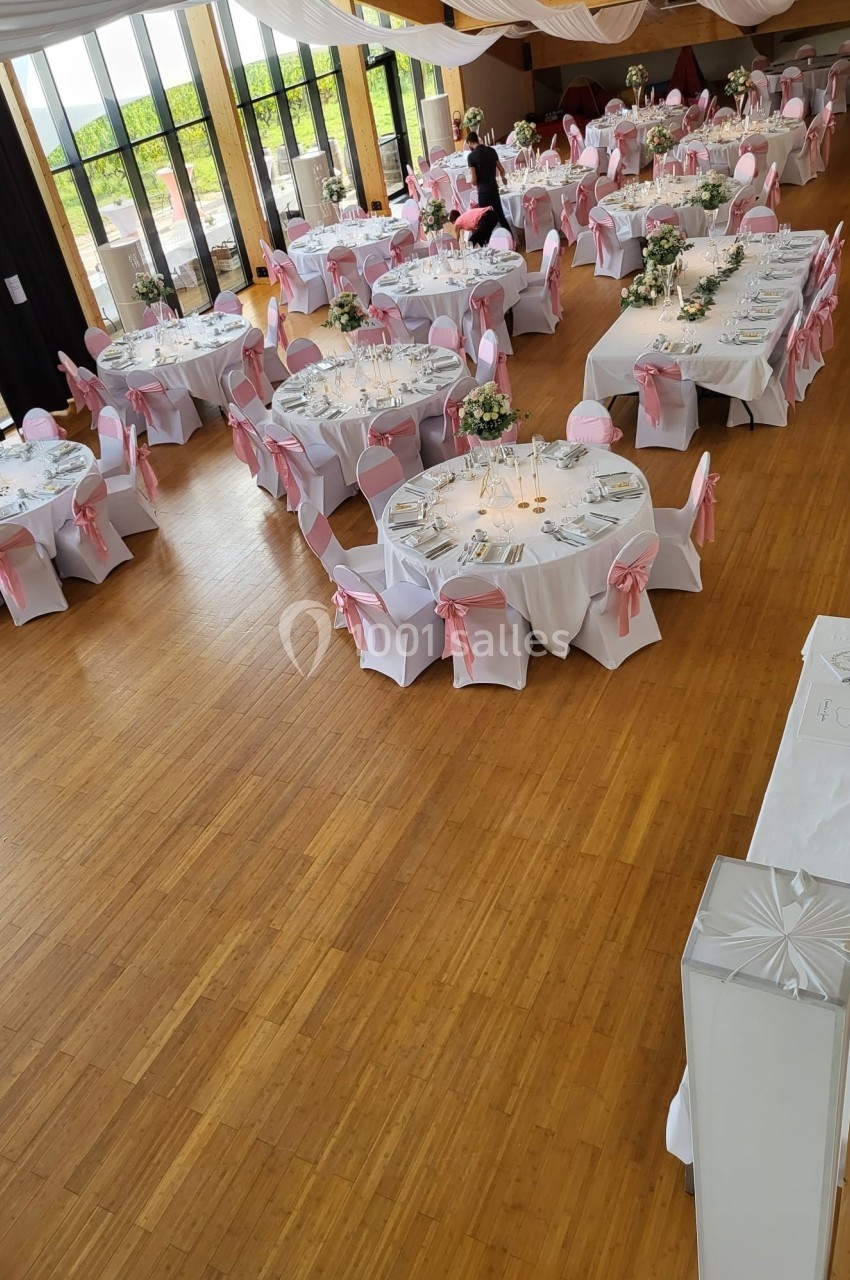 Salle de réception décorée avec des tables rondes, nappes blanches et chaises ornées de nœuds roses.