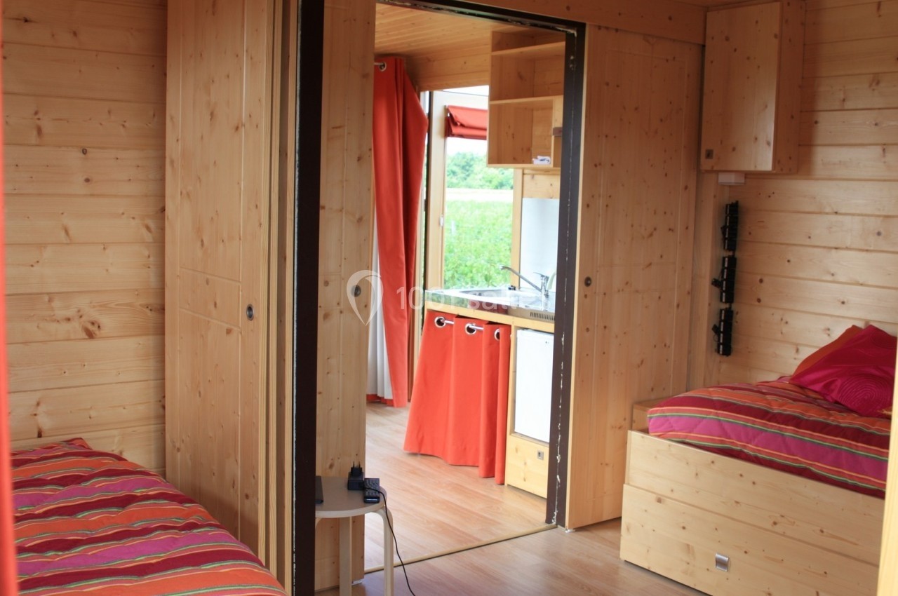 Intérieur d'un hébergement en bois avec deux lits, une kitchenette et des rideaux rouges donnant sur l'extérieur.