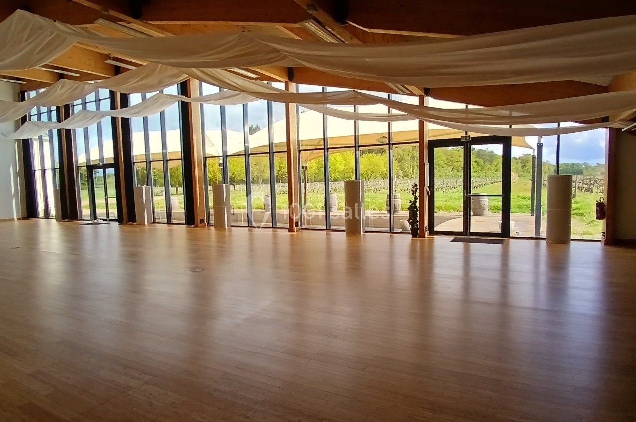 Salle lumineuse avec parquet en bois, grandes baies vitrées et rideaux blancs, donnant sur un espace extérieur verdoyant.