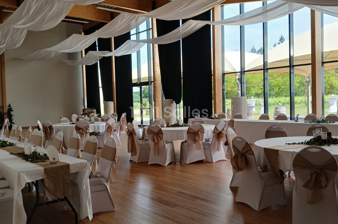 Salle de réception lumineuse avec tables rondes et rectangulaires décorées de nappes blanches et nœuds beige.