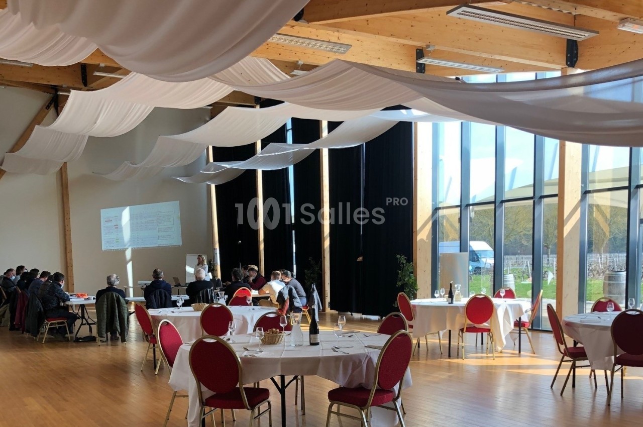 Salle lumineuse avec grandes baies vitrées, tables rondes dressées et participants réunis en groupe à gauche.