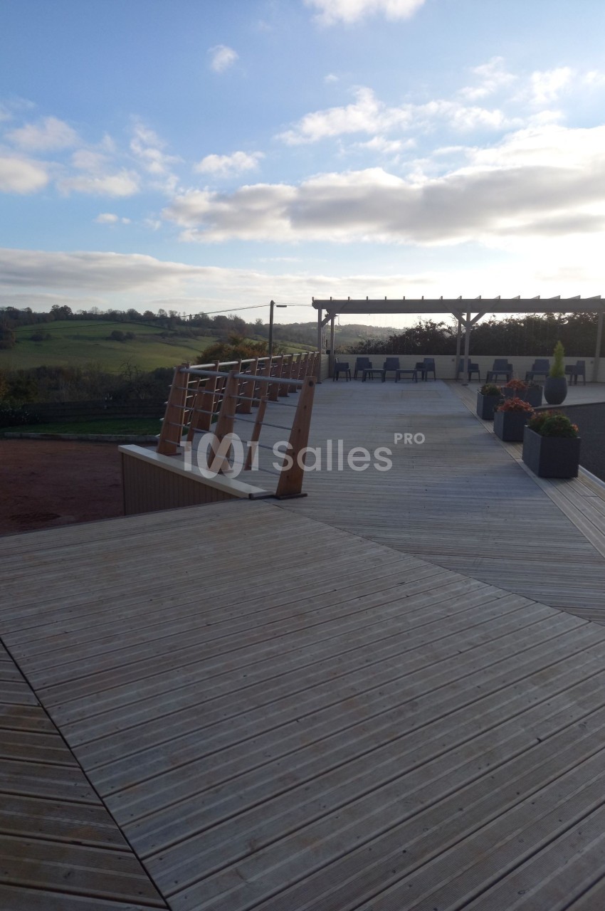 Terrasse en bois avec rampe d'accès, pergola et vue dégagée sur un paysage rural sous un ciel partiellement nuageux.