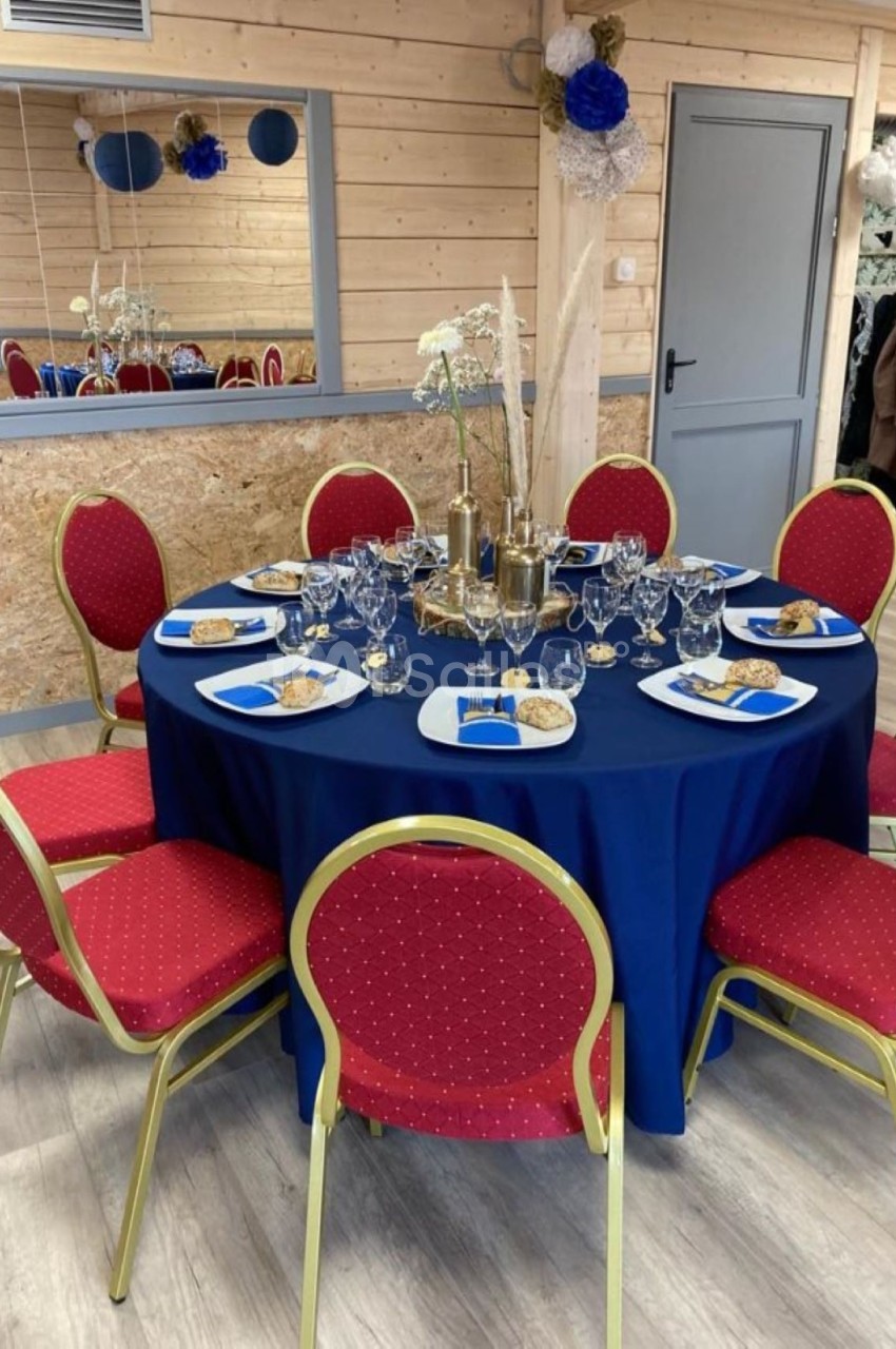 Table ronde dressée avec une nappe bleue, entourée de chaises rouges, décorée pour un repas avec vaisselle et ornements.