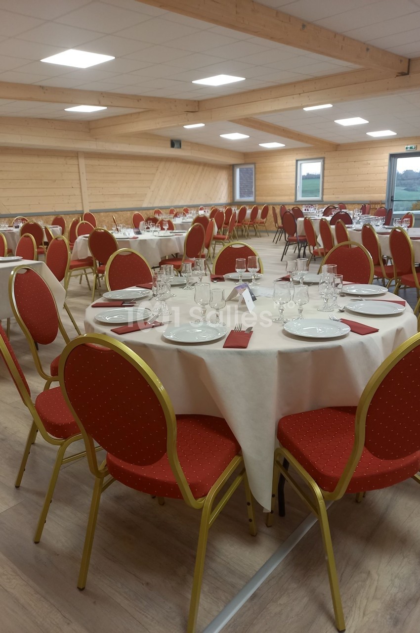 Salle de réception avec tables rondes dressées, nappes blanches, chaises rouges et couverts disposés pour un repas.