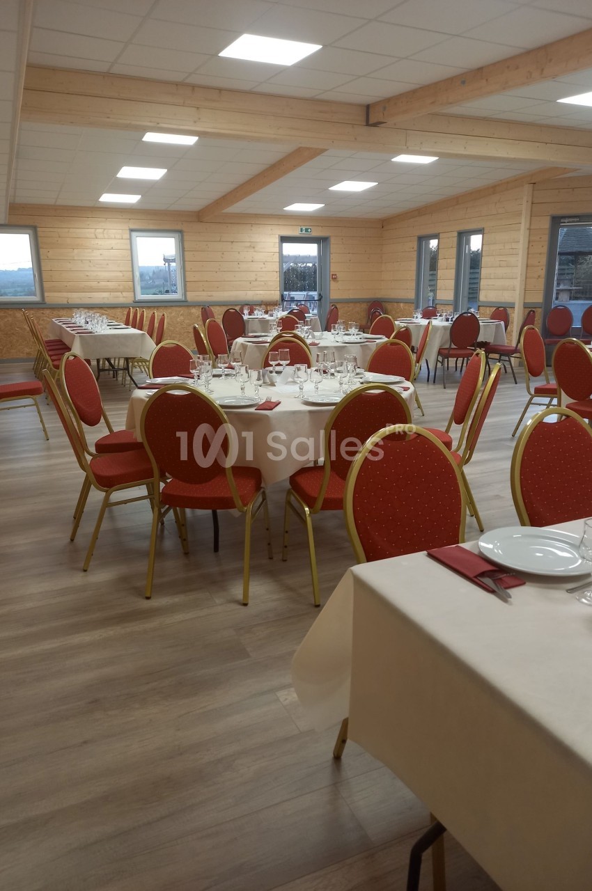 Salle de réception avec tables rondes dressées, nappes blanches, serviettes rouges et chaises rouges sur un sol en bois.