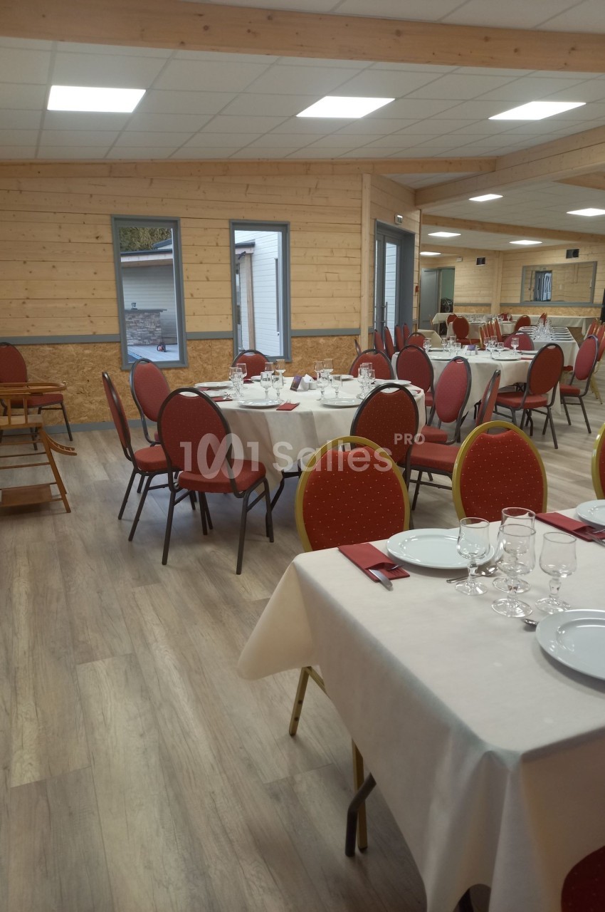 Salle de réception avec tables dressées, nappes blanches, chaises rouges et lumière naturelle provenant des fenêtres.