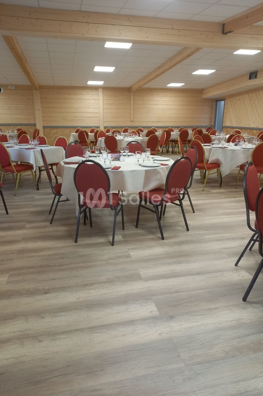 Salle de réception avec tables rondes dressées, nappes blanches et chaises rouges, dans un espace en bois clair.