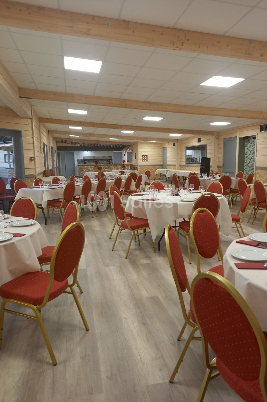 Salle de réception lumineuse avec tables rondes dressées, chaises rouges et décor en bois clair.