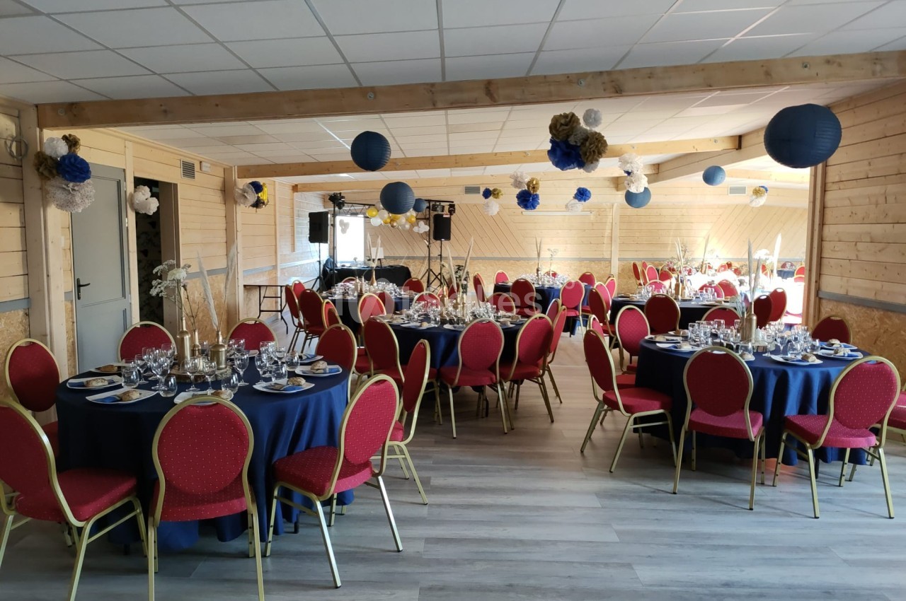 Salle décorée pour un événement, avec tables rondes dressées, nappes bleues, chaises rouges et décorations suspendues.