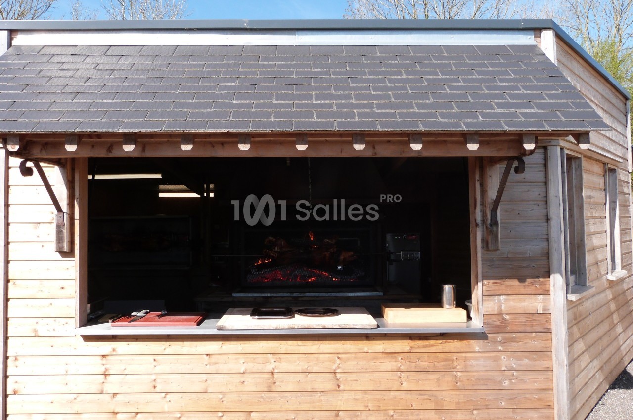 Abri en bois avec un four à rôtisserie visible à travers une ouverture, entouré d'arbres sous un ciel dégagé.