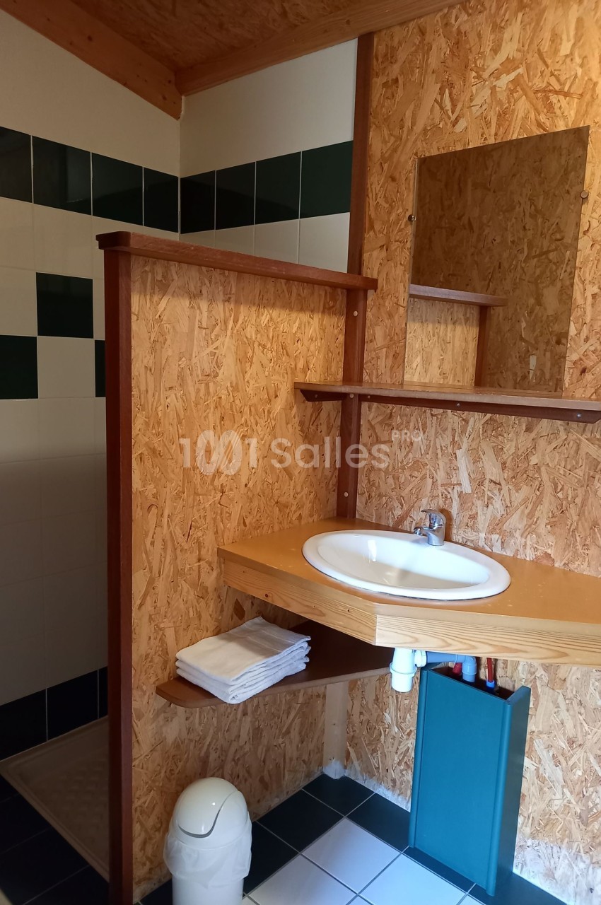 Salle de bain avec lavabo, miroir, étagères en bois, douche carrelée et poubelle blanche sur un sol en carrelage.