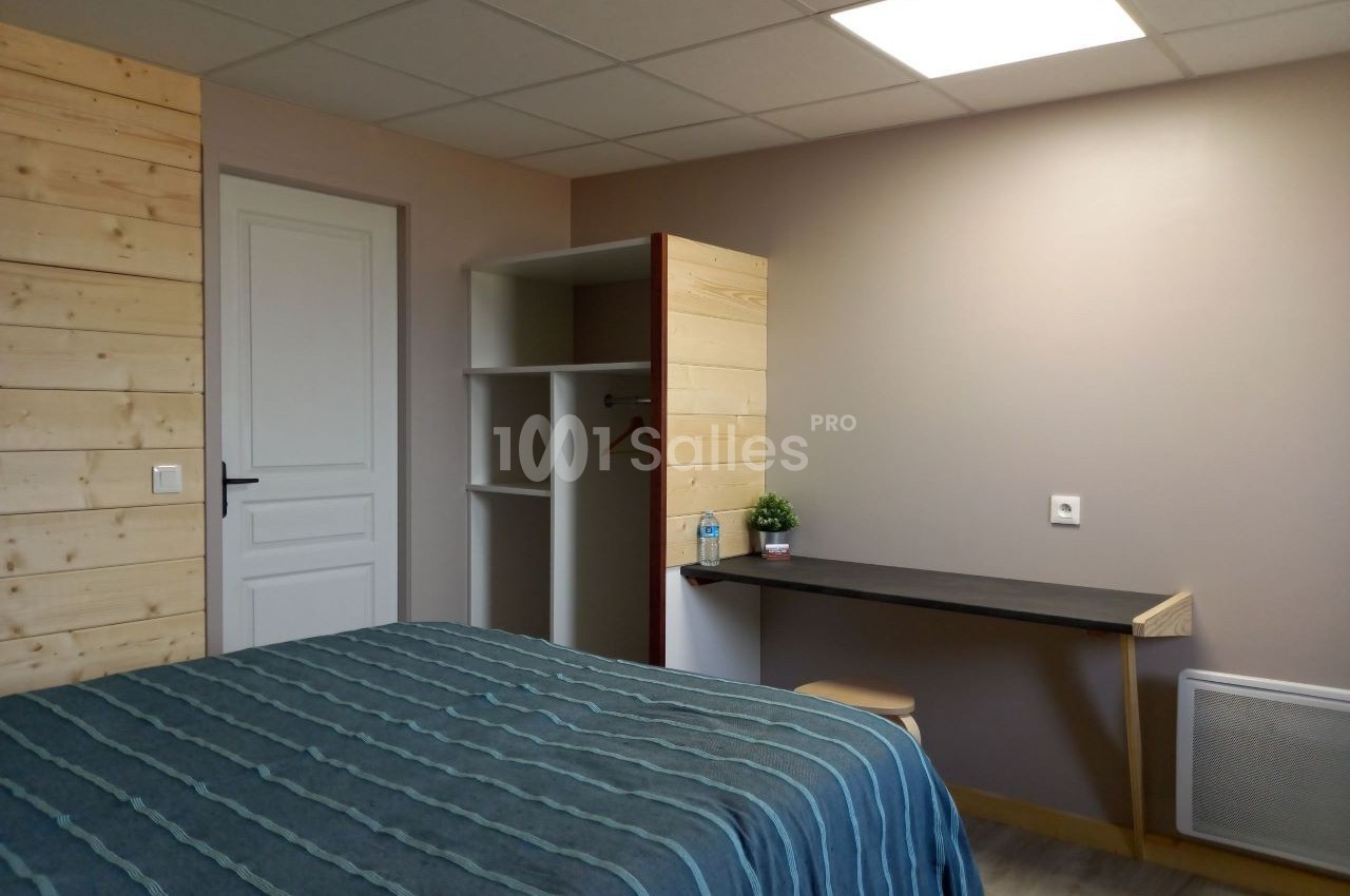 Chambre simple avec lit, bureau, étagères ouvertes et mur en bois, éclairée par une lumière au plafond.