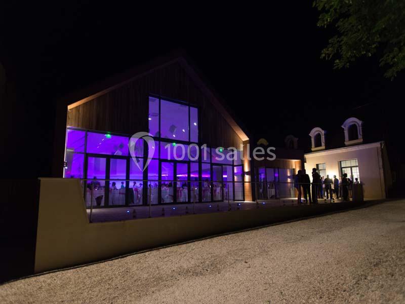 Bâtiment moderne avec façade vitrée illuminée en violet, vu de nuit, avec des personnes rassemblées à l'extérieur.