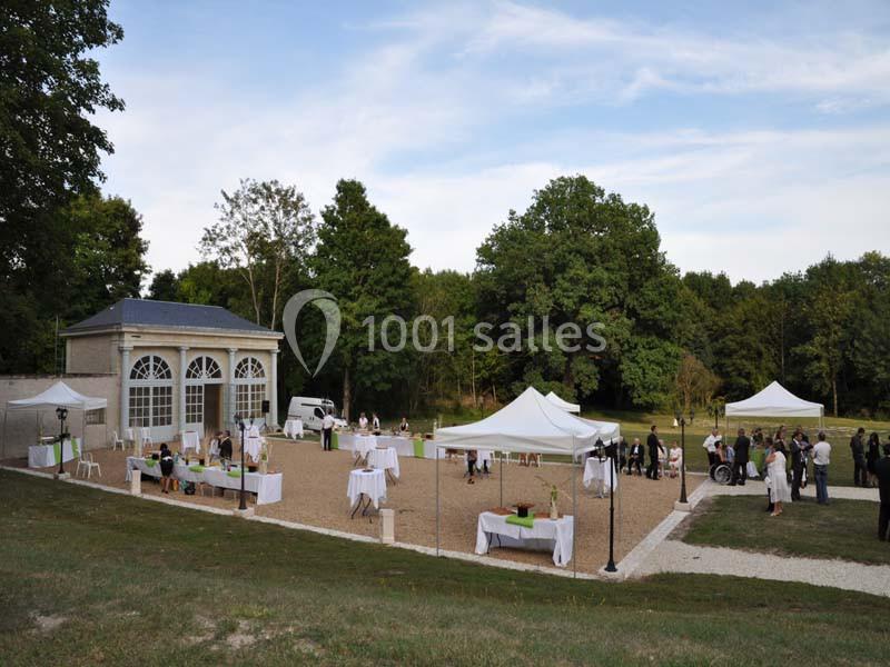 Jardin avec une orangerie, tentes blanches et tables dressées, entouré d'arbres, lors d'un événement en plein air.