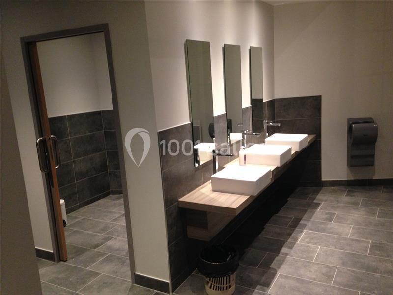 Salle de bain moderne avec trois lavabos, miroirs, carrelage gris et distributeur de papier mural.