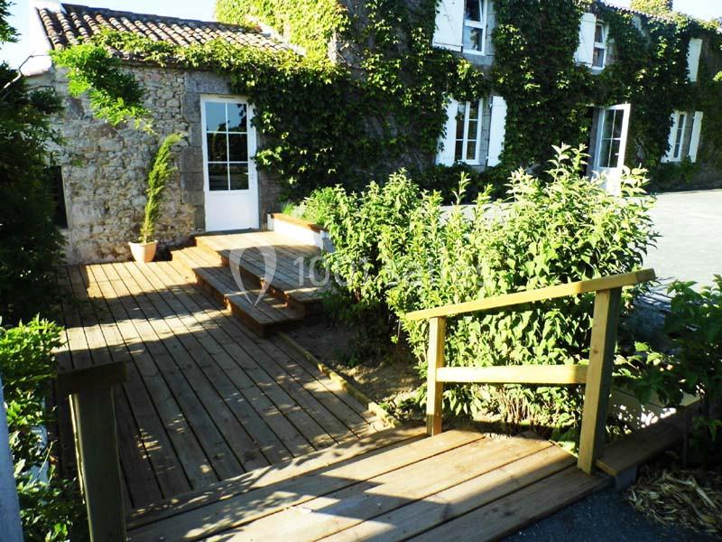 Terrasse en bois avec rampe et marches, devant une maison en pierre recouverte de lierre, par une journée ensoleillée.