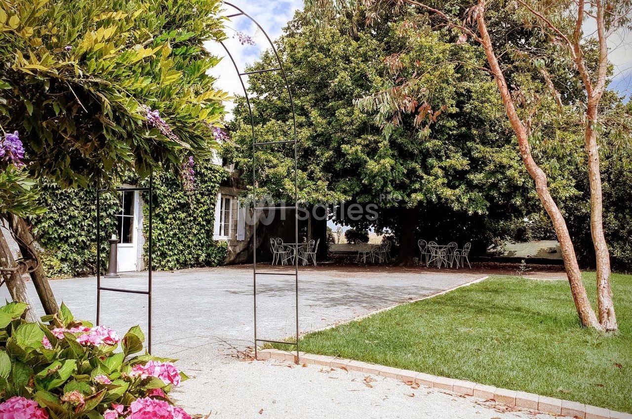 Jardin avec pelouse, terrasse pavée, mobilier de jardin sous des arbres et arche métallique entourée de fleurs.
