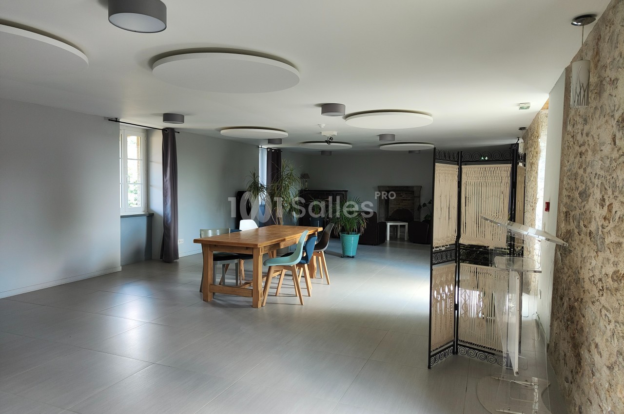 Salle lumineuse avec table en bois, chaises, plantes en pot, murs clairs et éléments en pierre apparente.