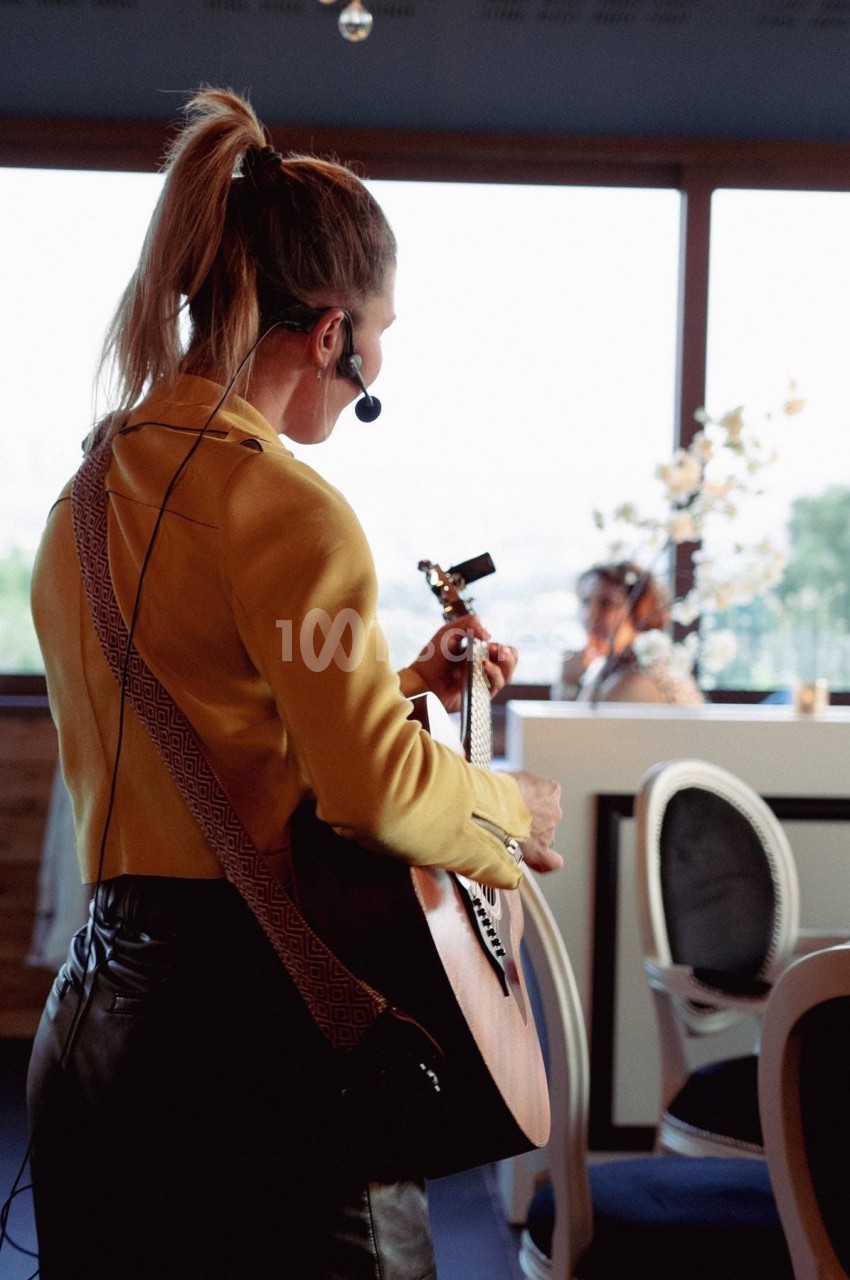 Une musicienne joue de la guitare avec un micro-casque dans un restaurant lumineux avec vue sur l'extérieur.