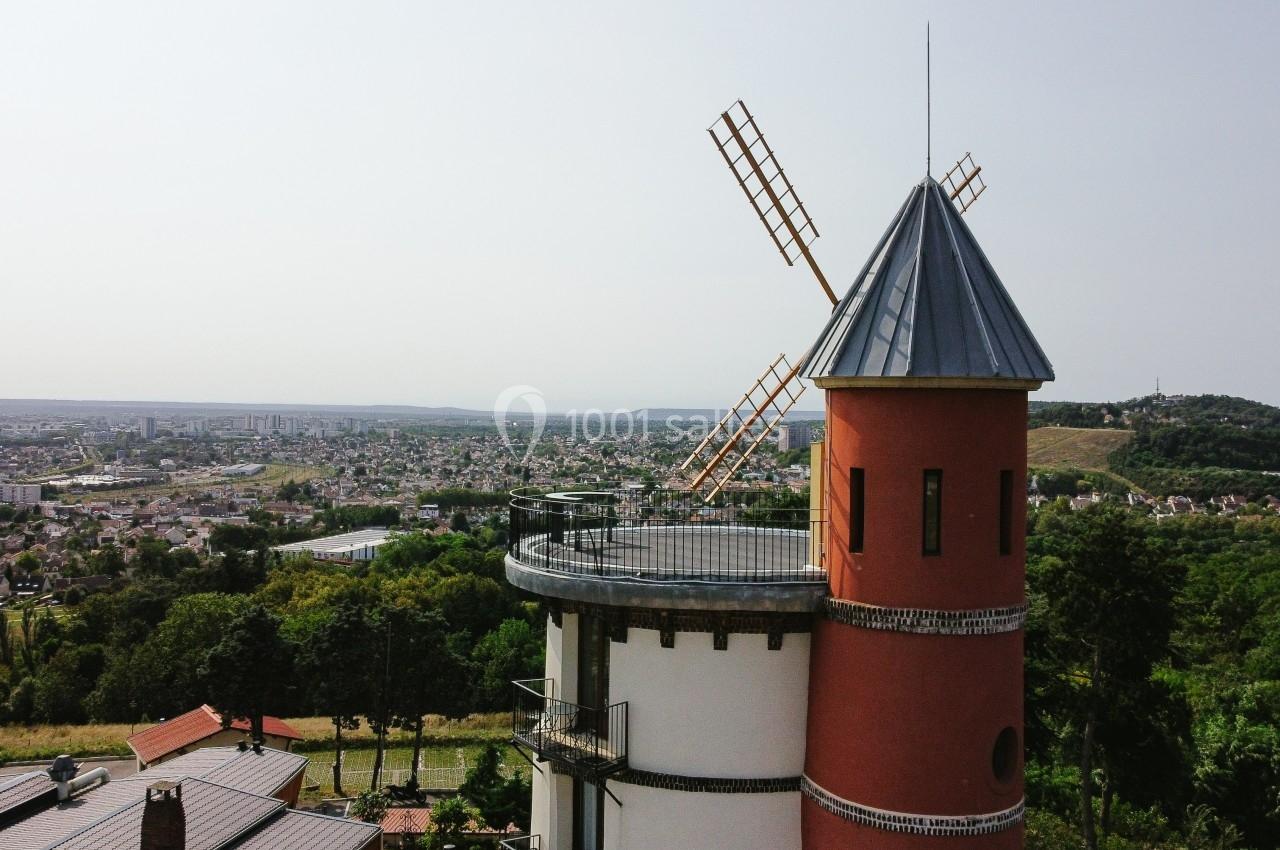 Vue d'un moulin à vent rouge avec toit en ardoise, surplombant une ville et des collines verdoyantes en arrière-plan.
