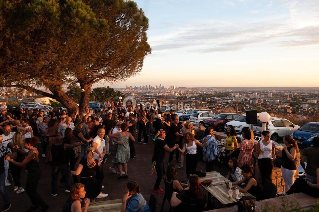 Groupe de personnes dansant et socialisant en extérieur au coucher du soleil, avec vue sur une ville en arrière-plan.
