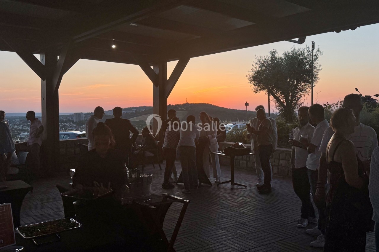 Groupe de personnes réunies sous une terrasse ouverte au coucher du soleil, avec vue sur un paysage vallonné.