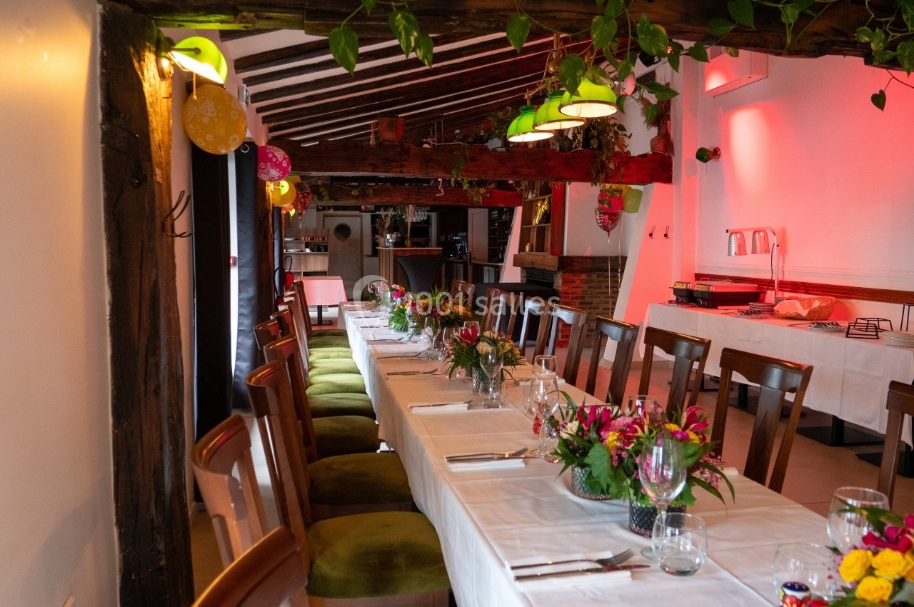Salle décorée pour un événement, avec une longue table dressée, des fleurs, des ballons et un éclairage chaleureux.