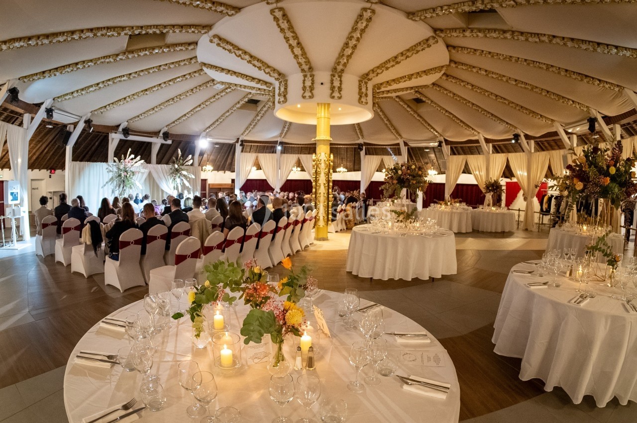 Salle de réception décorée avec des tables dressées, des chaises alignées et des fleurs, prête pour un événement.