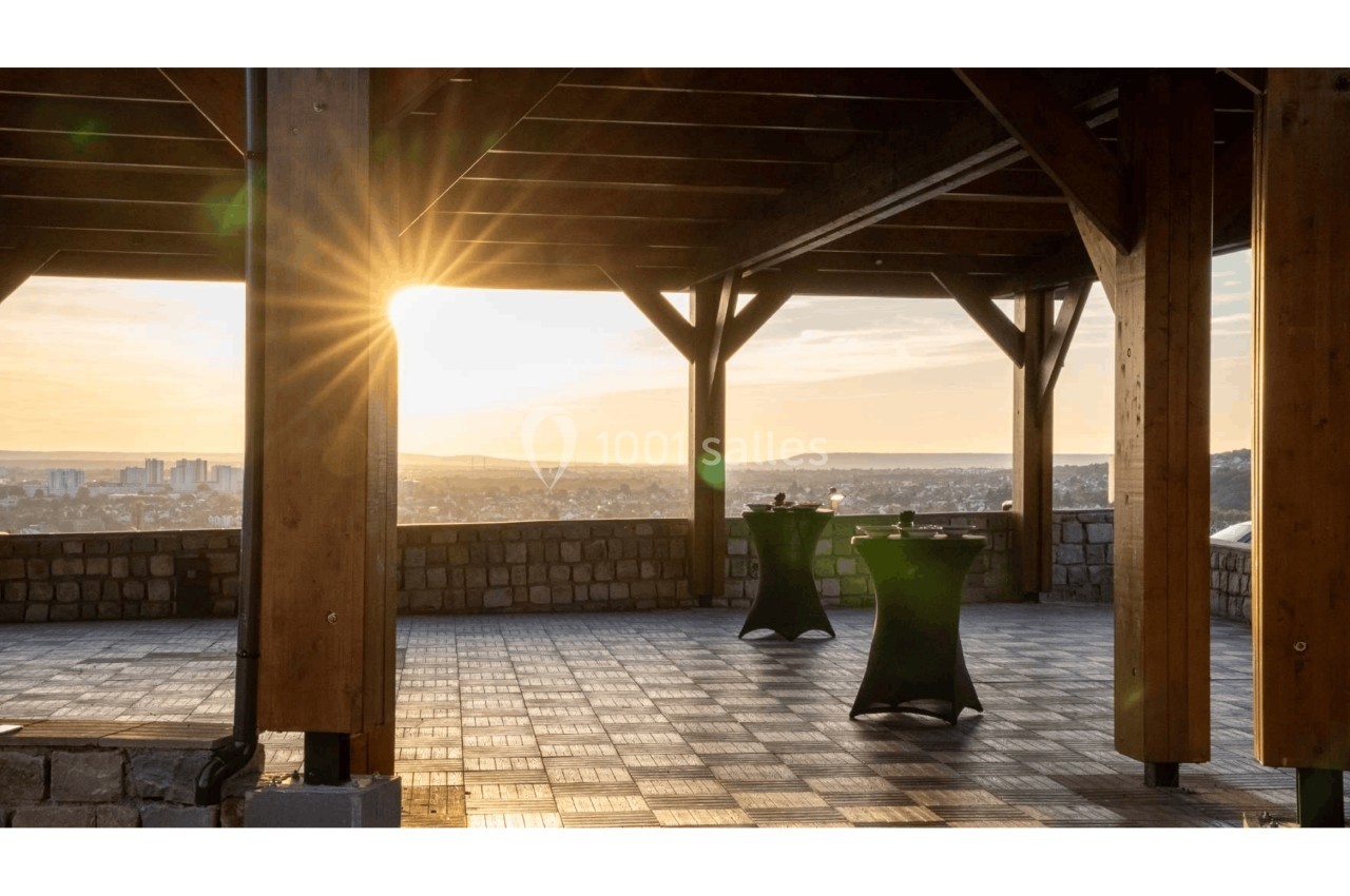 Vue d'une terrasse en bois avec des tables hautes, éclairée par un coucher de soleil surplombant une ville.