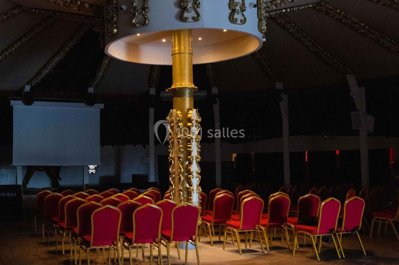 Salle de conférence sombre avec des rangées de chaises rouges et une colonne dorée centrale ornée de motifs.
