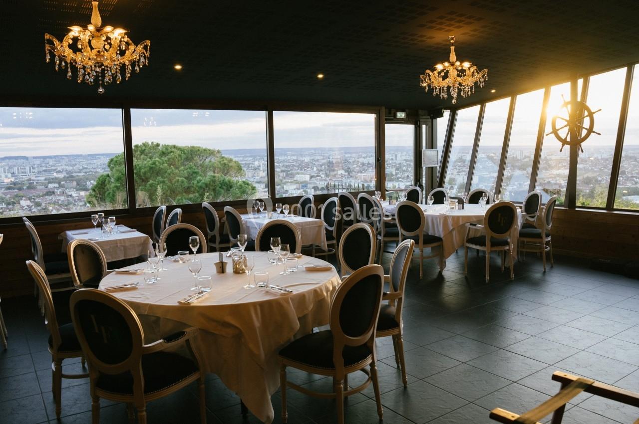 Salle de restaurant élégante avec grandes baies vitrées offrant une vue panoramique sur une ville au coucher du soleil.
