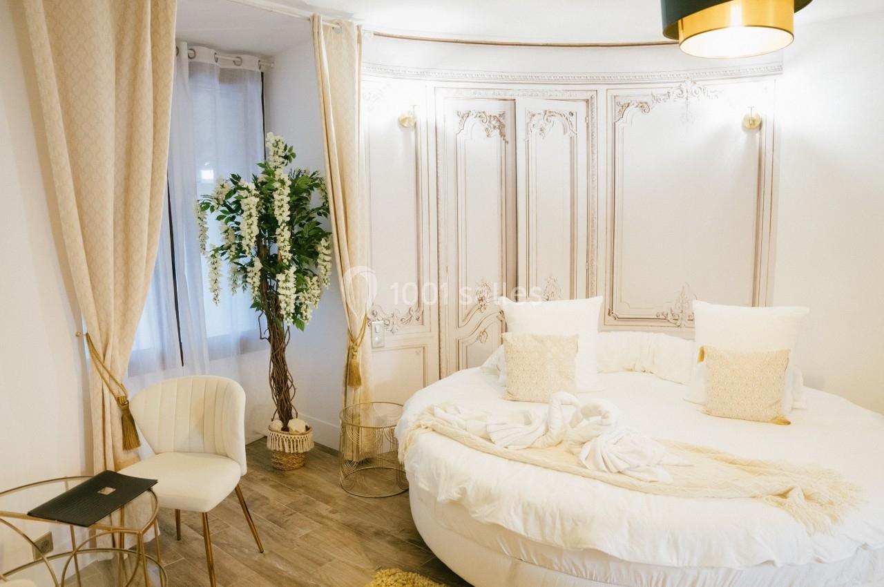 Chambre lumineuse avec lit rond, décor élégant, rideaux beiges, plante verte et mobilier doré sur parquet clair.