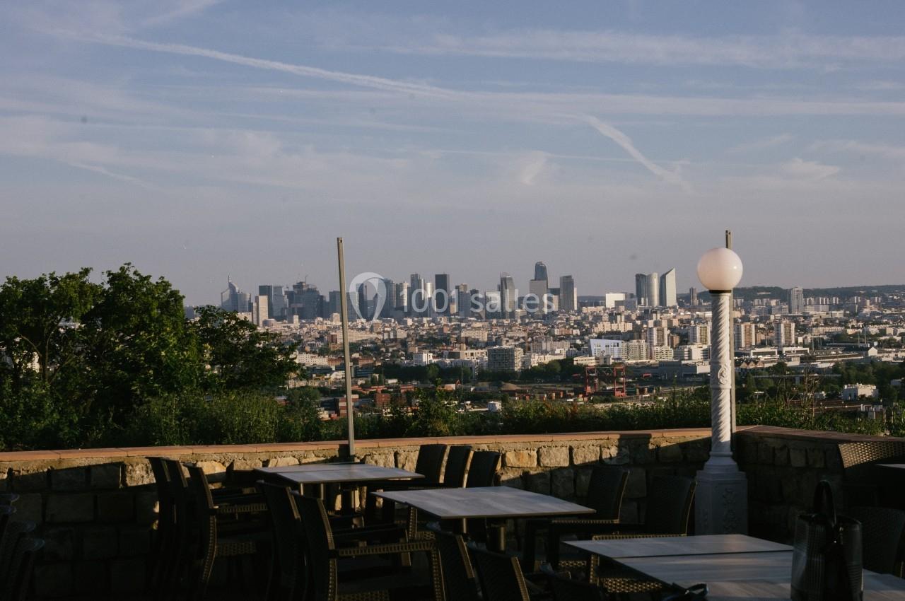 Vue d'une terrasse avec des tables et des chaises, donnant sur une ville et des gratte-ciel à l'horizon sous un ciel dégagé.