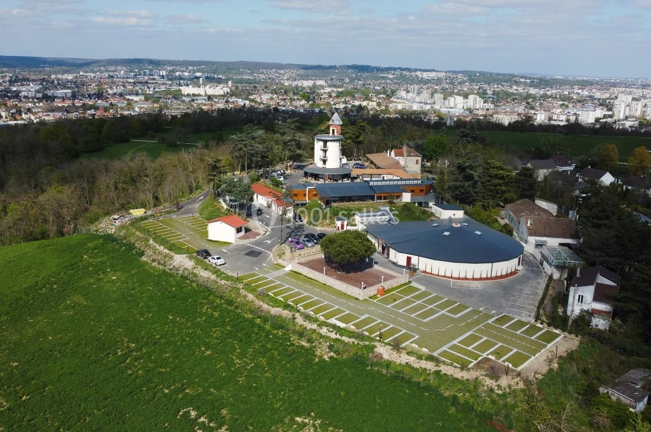 Vue aérienne d'un bâtiment avec une tour blanche entouré de verdure, situé en périphérie d'une ville.