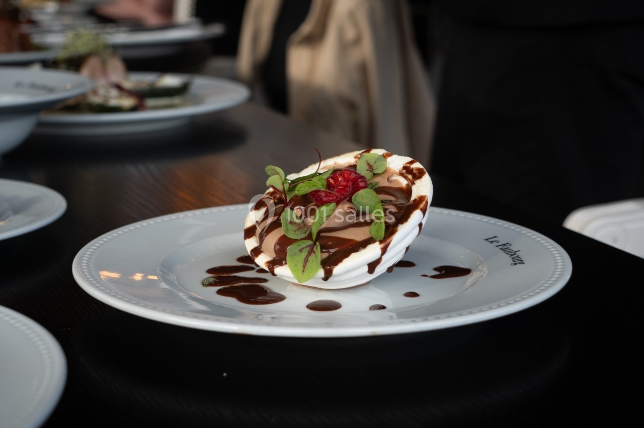 Dessert dressé sur une assiette blanche, garni de crème, chocolat, framboise et feuilles vertes, avec sauce chocolatée.
