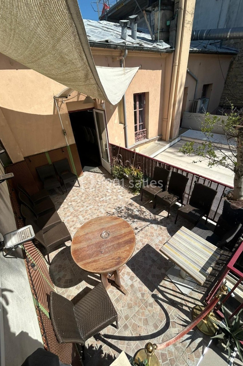 Terrasse ensoleillée avec table ronde en bois, chaises en rotin, plantes et voiles d'ombrage dans un cadre urbain.
