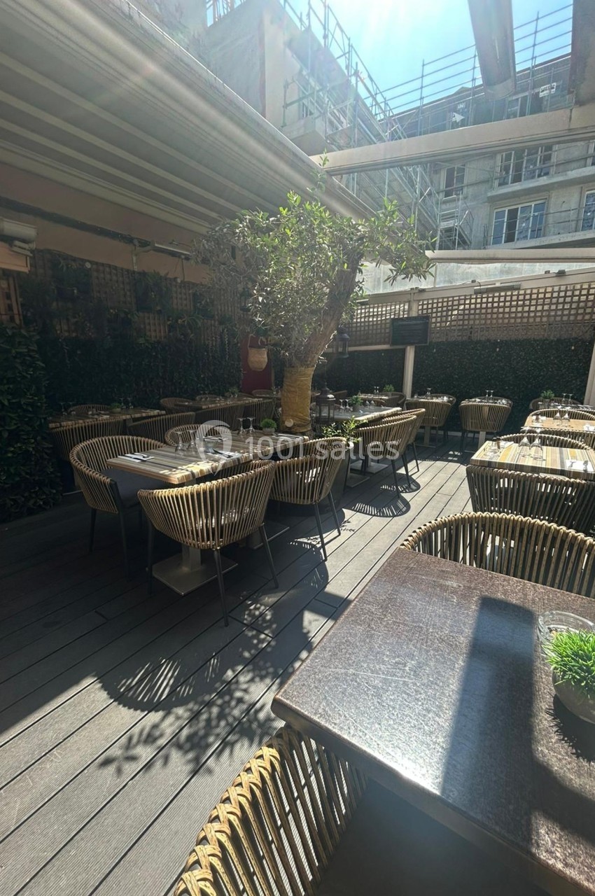 Terrasse extérieure d'un restaurant avec des tables dressées, des chaises en rotin et un grand arbre au centre.