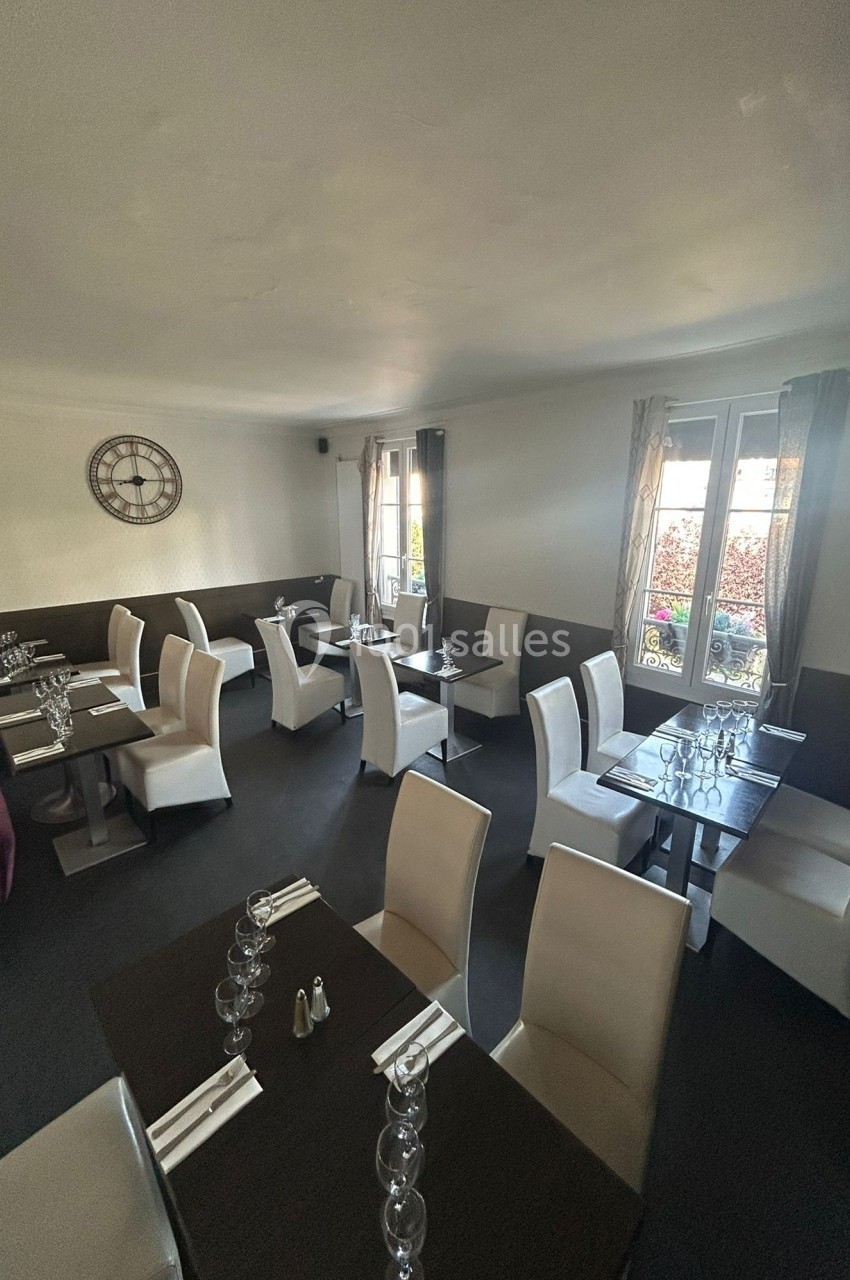 Salle de restaurant lumineuse avec des tables dressées, chaises blanches et une horloge murale décorative.
