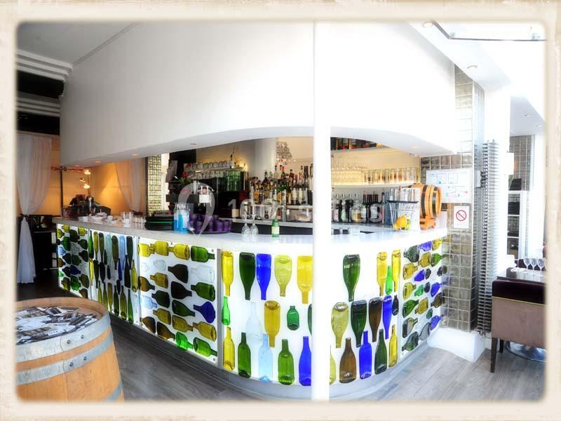 Bar lumineux avec comptoir décoré de bouteilles colorées, entouré de tabourets et d'étagères garnies de bouteilles.