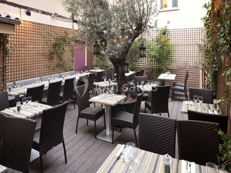 Terrasse extérieure avec des tables dressées, des chaises en osier, un grand olivier central et des panneaux en bois ajourés.