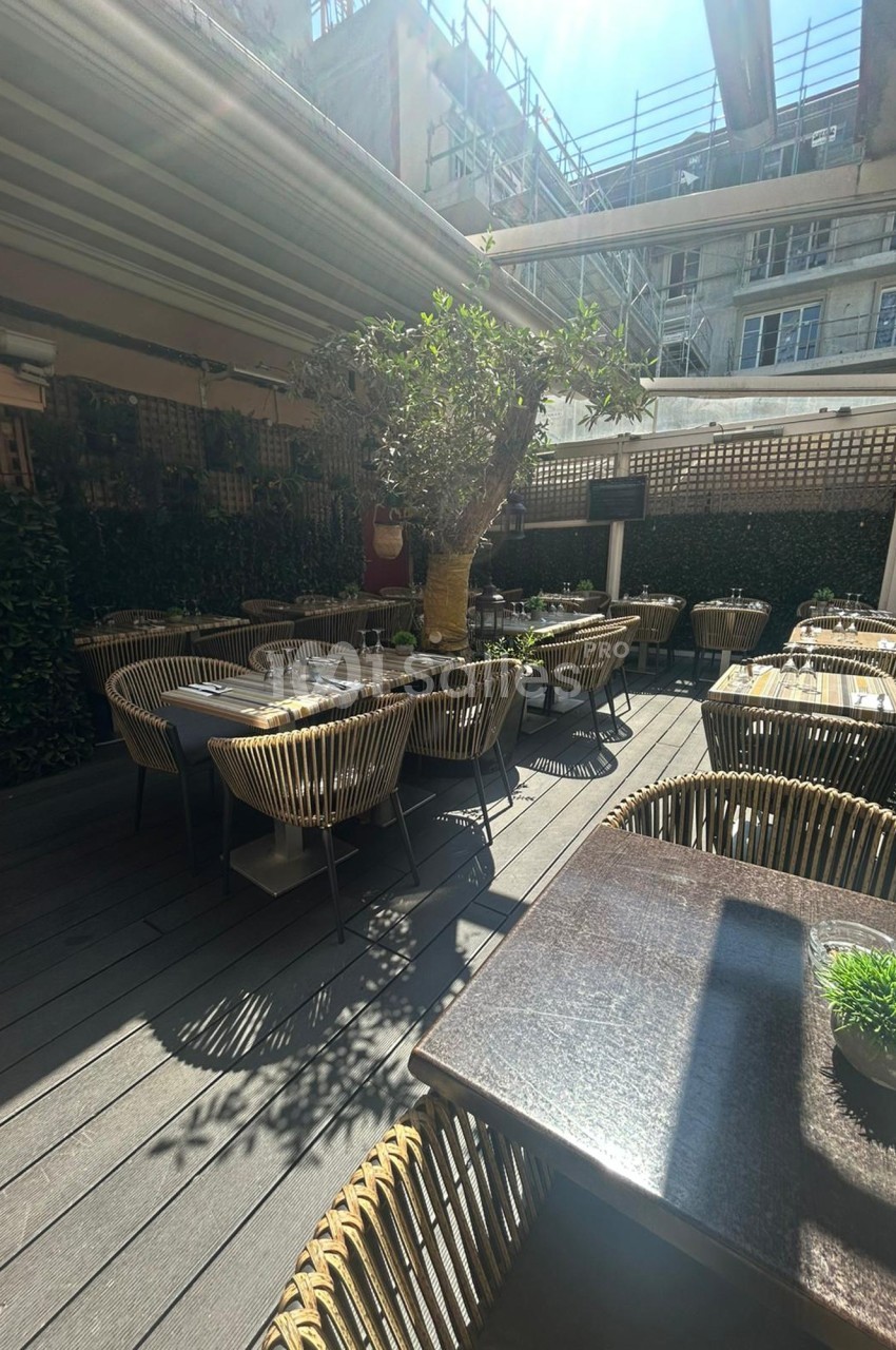 Terrasse extérieure d'un restaurant avec des tables dressées, des chaises en rotin et un grand arbre au centre.