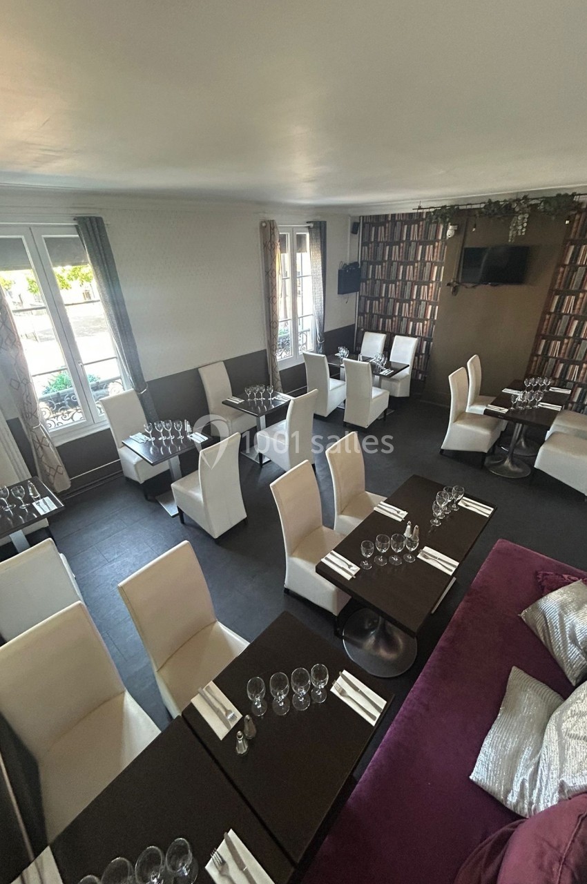 Salle de restaurant élégante avec tables dressées, chaises blanches et décoration murale imitant une bibliothèque.
