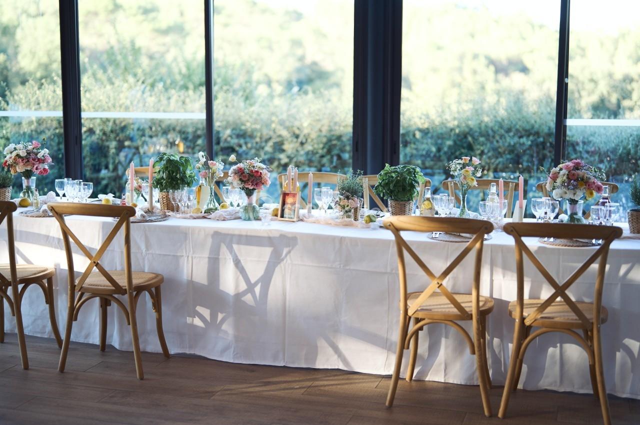 Table décorée pour un repas, avec nappes blanches, fleurs, vaisselle et chaises en bois, près de grandes baies vitrées.