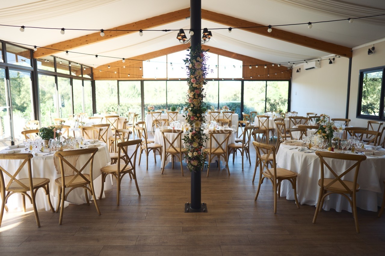 Salle de réception lumineuse avec tables rondes dressées, chaises en bois et décoration florale sur une colonne centrale.